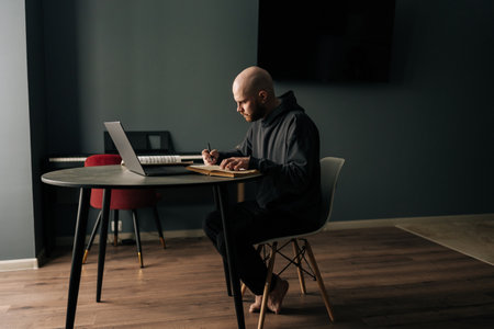 Remote side view of focused freelancer male taking notes sitting at table with laptop computer, managing online business and enjoying comfort of working from home. Concept of remote job, technology.の写真素材