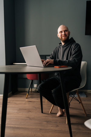 Full length vertical portrait of cheerful programmer male remote working from home office, sitting at wooden table, typing on laptop computer looking away with happy expression.の写真素材