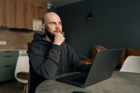 Portrait of pensive freelancer male sitting at table, working on laptop computer while resting hand on chin, deep in thought looking away, working remotely from cozy home office environment.の写真素材