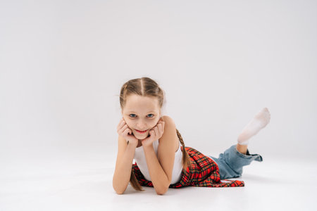 Studio portrait of charming schoolgirl with braided hair wearing white tank top, red plaid shirt and jeans, lying on white background, resting chin on hands, smiling looking at camera.の写真素材
