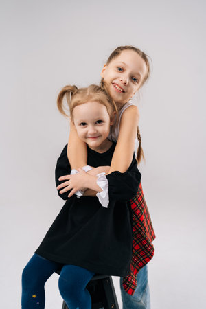 Vertical portrait of two young sisters embracing and smiling looking at camera in studio with white isolated background, radiating love and joy, capturing of close bond and carefree childhood.の写真素材