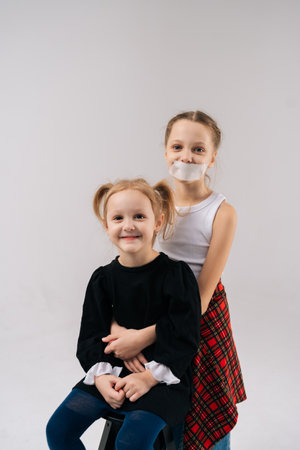 Studio portrait of two young sisters, one with mouth covered by tape, embracing each other against isolated white background.の写真素材