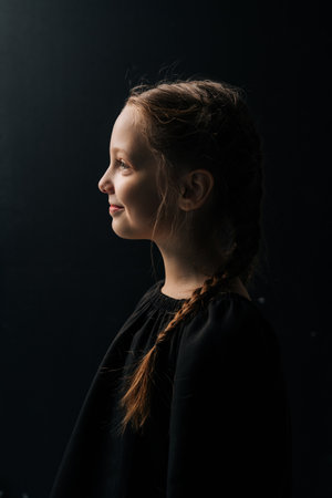 Side view portrait of charming tween girl with braided hair, wearing black dress, posing against dark isolated background in studio. Close-up face of adorable child illuminated by soft light.の写真素材