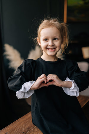 Vertical portrait of charming 5 years old girl with blond hair and black dress making heart shape with hands sitting on wooden bench in cozy indoor setting.の写真素材