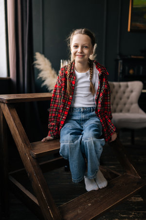 Full length vertical portrait of smiling preteen with braided hair wearing plaid shirt, sitting on ladder against dark wall. Cheerful child girl posting in casual clothes smiling looking at camera.の写真素材