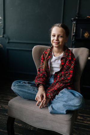Vertical portrait of pretty preteen girl with braided hair wearing casual clothes sitting cross-legged on armchair in dark room, looking serene and comfortable. Adorable child posing at home alone.の写真素材
