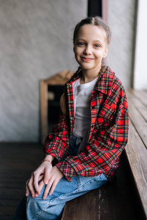 Vertical portrait of cheerful tween girl with braided hair wearing red plaid shirt and jeans, sitting on bench by window, enjoying relaxed moment, smiling looking at camera with happy expression.の写真素材