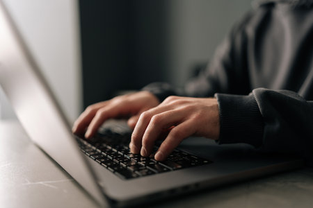 Close-up hands of unrecognizable male typing on laptop keyboard, immersed in software development or coding, sitting at table by window in dark room. Concept of remote job, freelance.の写真素材