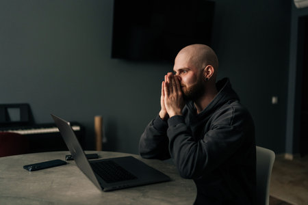 Side view of exhausted freelancer male covering face with hand while sitting at desk in home office, feeling tired and overwhelmed by work. Concept of remote job, technology and people.の写真素材