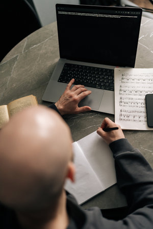 Top view from shoulder of unrecognizable composer male creating music notes on paper while using laptop, surrounded by sheet music, book, smartphone on marble table in cozy home workspace.の写真素材