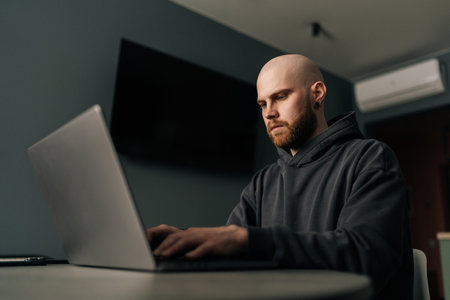 Concentrated male programmer in casual clothes typing code on laptop, developing software in dark...