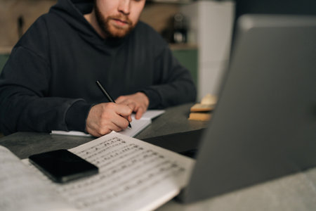 Cropped shot of music composer writing melodic notes in home studio, sitting at table with laptop, smartphone and sheet music scattered across workspace, merging artistic expression with digital toolsの写真素材
