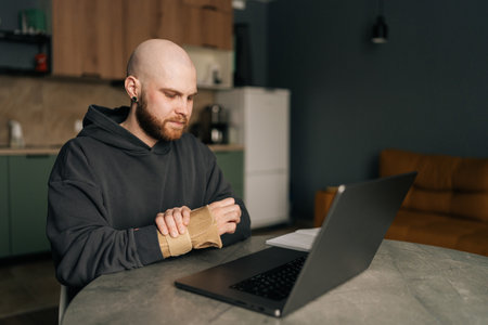 Sick freelancer programmer with wrist brace rubbing painful hand, working on laptop during carpal tunnel syndrome management at home workspace, sitting at table on background of kitchen interior.の写真素材