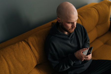 Top view of focused young bald man with beard and hoodie using smartphone while sitting on comfortable sofa, browsing internet, social media or mobile application. Concept of modern technology.の写真素材
