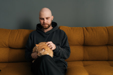 Portrait of young bald man with beard wearing casual clothes sitting on comfortable sofa, lovingly petting ginger cat, enjoying quiet moment at home. Concept of relationship between people and animal.の写真素材