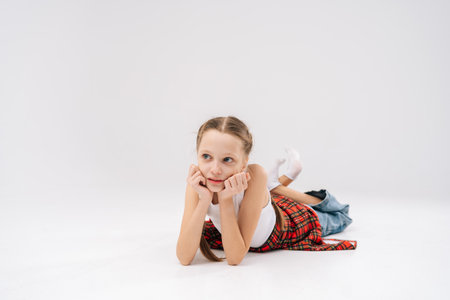 Pretty 10 years old girl lying on floor, chin propped on hands, sporting white tank and red checkered shirt, exuding relaxed mood in studio with isolated background. Concept of carefree childhood.の写真素材