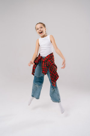 Cheerful 10 years old girl jumping mid-air with wide smile in studio with white isolated background. Happy preteen grinning widely, sporting white tank top, plaid shirt knotted at waist, baggy jeans.の写真素材