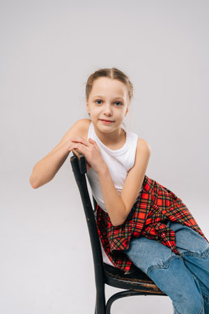 Blue-eyed preteen with light brown hair wearing white tank top, red checkered shirt and jeans, sitting posing on vintage black chair in studio with white isolated background, looking at camera.の写真素材