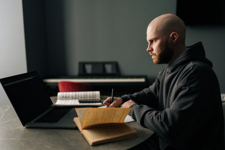 Focused talented composer writing music notes on paper sitting at table with laptop, books, sheet music in home office, immersing in creativity and inspiration. Concept of remote job, technology.の写真素材