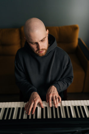 Vertical portrait of focused bearded male composer playing synthesizer in living room. Musician wearing dark hoodie creating melodic composition using digital piano at home studio.の写真素材