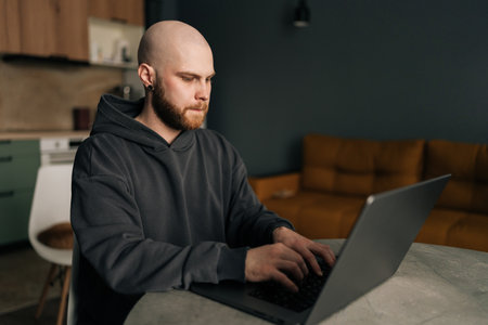 Focused programmer wearing dark hoodie typing on laptop computer, sitting at table near bright window in contemporary urban apartment with minimalist design and soft daylight.の写真素材