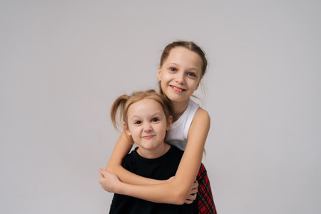 Adorable playful young sisters posing for studio portrait, with older sister affectionately hugging younger sister from behind, both smiling happily looking at camera. Concept of happy sisterhood.の写真素材