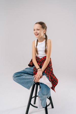 Cheerful preteen girl with two braids, wearing white sleeveless shirt, jeans, red checkered shirt tied around waist, sitting on chair in studio with white isolated background, smiling looking awayの写真素材