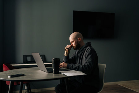 Focused freelancer with injured hand using laptop and drinking coffee while working from home office, sitting at desk with modern interior design, in cozy and productive workspace, looking to screen.の写真素材