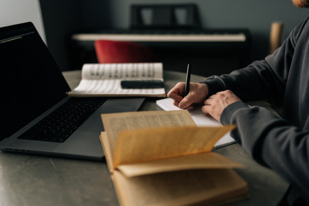 Close-up hands of unrecognizable composer writing music notes on paper sitting at table with laptop, books, sheet music in home office, immersing in creativity and inspiration. Concept of remote job.の写真素材