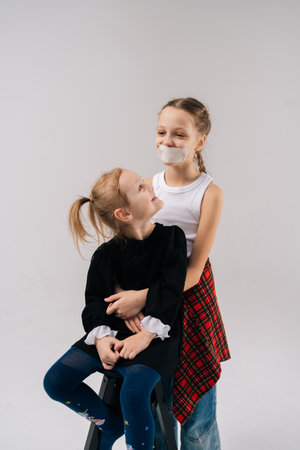 Compelling studio portrait showing young sisters hugging closely, one sibling wearing tape across mouth, symbolizing silenced voices on isolated white background. Concept of sisterhood.の写真素材