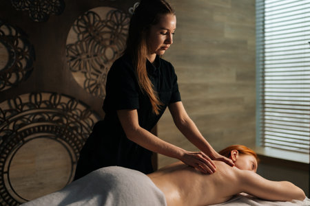 Professional massage therapist performing relaxing back massage for female client on treatment table, on background of wall in tranquil wellness environment. Concept of healthy lifestyleの写真素材