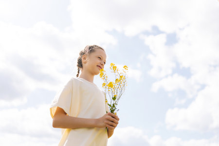 Low-angle view of happy young girl with braided hair holding bouquet of yellow wildflowers standing posing in sunny summer meadow, enjoying beauty of nature under bright cloudy sky.の写真素材