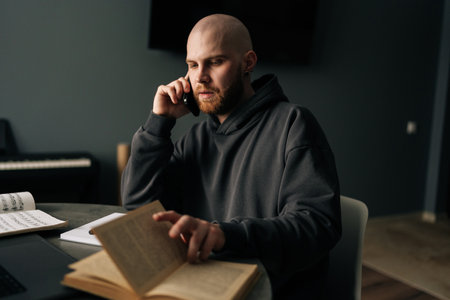 Portrait of talented music composer talking on phone and reading book at home studio, with laptop and sheet music on table, working remotely on new project. Concept of remote job, technology, people.の写真素材