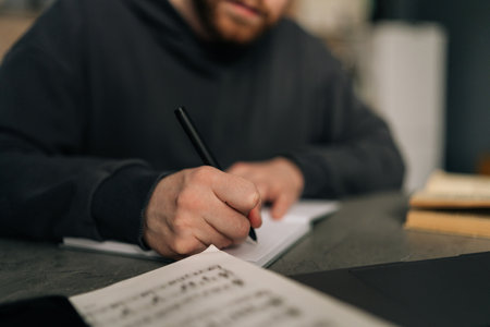 Close-up of music composer male writing melodic notes in home studio, sitting at table with laptop and sheet music scattered across workspace, merging artistic expression with digital toolsの写真素材