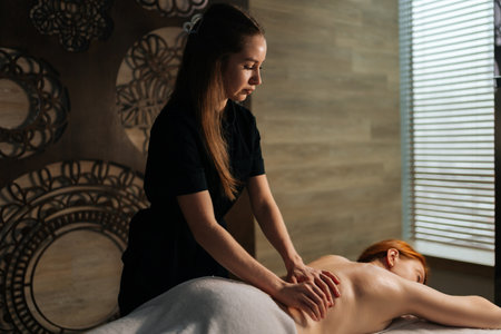 Focused professional masseuse performing therapeutic back massage to young woman lying resting on massage table in serene spa environment, enhancing wellness and promoting deep relaxationの写真素材