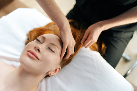 Close-up top view of skilled masseuse gently massaging temples of relaxed redhead woman with closed eyes, providing soothing head massage on comfortable massage table in spa or wellness center.の写真素材