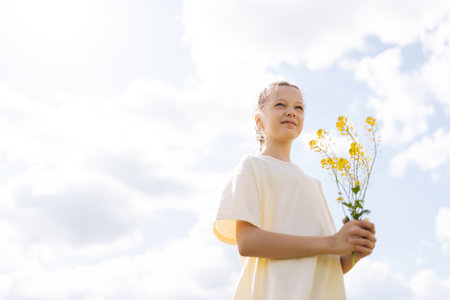 Adorable girl with braided hair holding yellow wildflowers, standing in sunlit summer meadow with cloudy sky in background, enjoying beautiful nature. Concept of happy childhood and outdoor recreationの写真素材