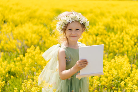 Cheerful 5 yo girl wearing flower crown and fairy wings holding blank canvas on wooden easel standing posing in field of yellow flowers, enjoying sunny summer day of artistic inspiration.の写真素材