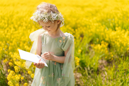 Portrait of adorable child girl wearing flower crown and green dress, painting on canvas, standing at picturesque field of blooming yellow flowers on sunny summer day, warm weather.の写真素材