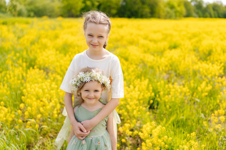 Portrait of adorable loving preteen and preschooler sibling standing hugging tightly in vibrant field of yellow flowers, enjoying sunny summer day together, looking at camera with happy expression.の写真素材