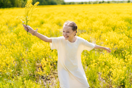 Cheerful young girl wearing light dress running and spinning through field of yellow flowers on sunny summer day, holding bouquet of wildflowers and enjoying nature. Concept of happy childhoodの写真素材