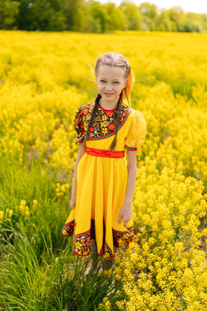 Vertical portrait of cute child girl in yellow dress with traditional Russian pattern, standing posing in field of yellow flowers during sunny summer day, enjoying beauty of nature and warm weather.の写真素材