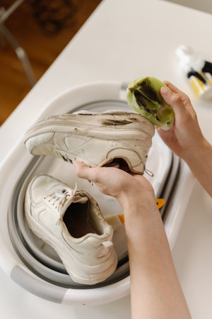 Top view of unrecognizable young man washing dirty white sneakers with cleaning foam and brush in basin, applying detergent on footwear, doing housework and chores, close-up.の写真素材