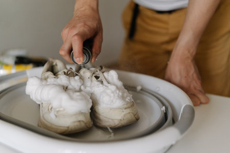 Close-up hands of unrecognizable man spraying cleansing foam on dirty white sneakers in basin, engaging in shoe cleaning and care within comfort of home, ensuring footwear hygiene and maintenance.の写真素材