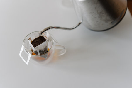 Top view of pouring hot water from kettle into drip coffee bag nestled in glass cup, brewing fresh cup of coffee on white table, close-up. Process of making brewed coffee at home during breakfast.の写真素材