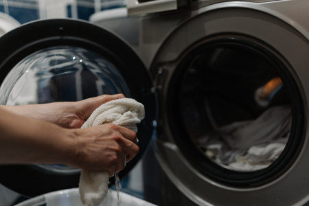 Close-up cropped shot of unrecognizable man wringing wet white cloth near washing machine. Housekeeper squeezing wet white cloth after washing in washing machine in modern laundry room.の写真素材