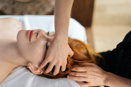 Close-up top view of professional masseuse massaging temples of young relaxed woman lying with closed eyes on massage table in spa salon. Masseur performing head massage for resting female, closeup.の写真素材