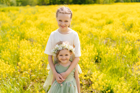 Loving preteen and preschooler sisters in flower crown standing hugging tightly in vibrant field of yellow flowers, enjoying sunny summer day together, looking at camera with happy expression.の写真素材