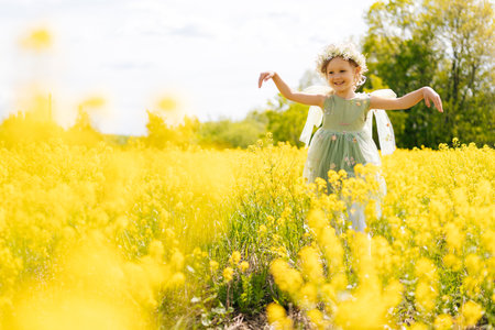 Happy preschooler girl wearing vibrant flower crown and fairy green dress, joyfully playing in sunny field filled with blooming yellow flowers in bright summer day. Concept of happiness childhoodの写真素材