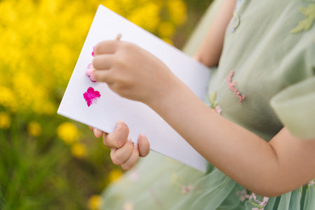 Close-up cropped shot of unrecognizable child girl wearing green dress painting small pink flowers on white canvas while standing in vibrant yellow meadow on sunny summer day enjoying beautiful natureの写真素材